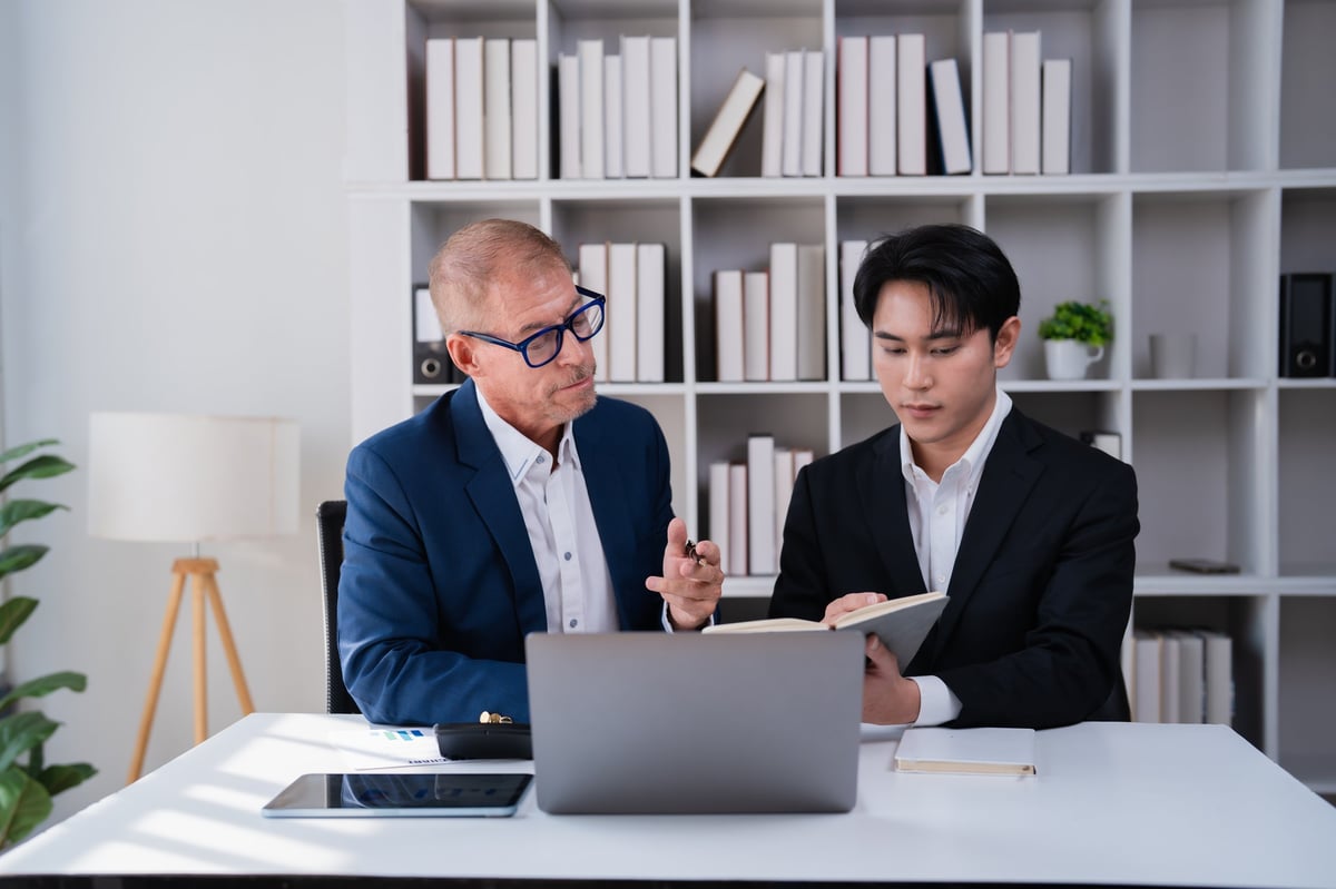 Two colleagues collaborate in a modern office, reviewing laptop data and notes while discussing strategy and mentoring across generations to solve business challenges together